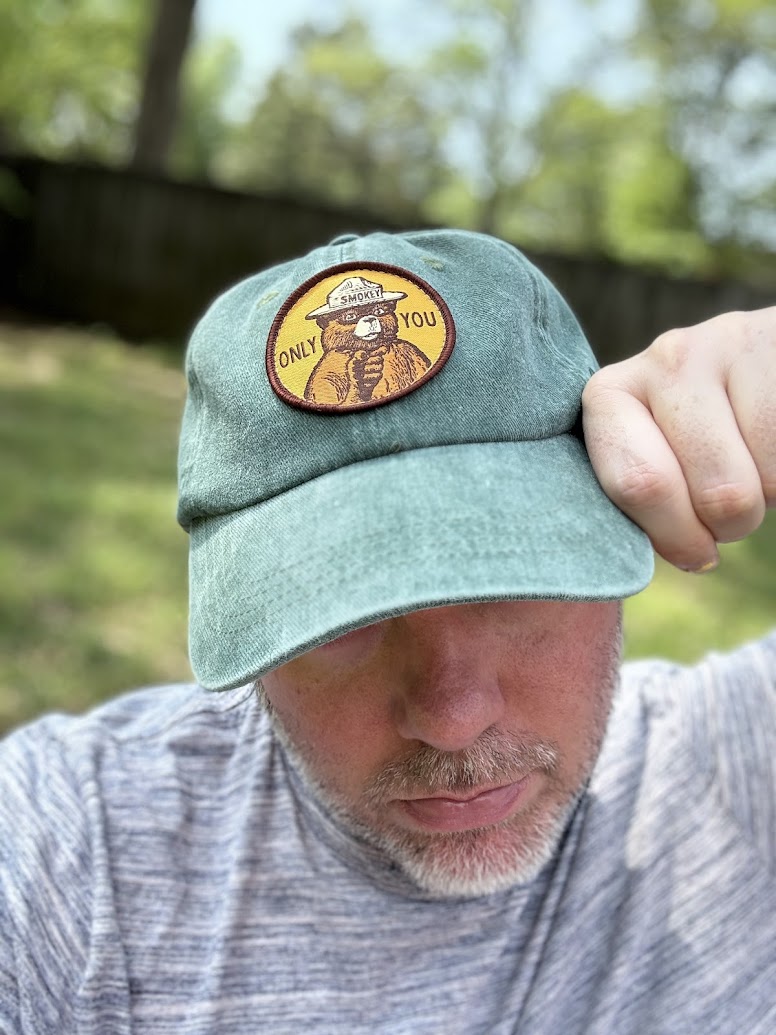 A person wearing a green cap with a Smokey Bear patch that reads "Only You." The person is looking down, and the background is a blurred outdoor setting with trees.