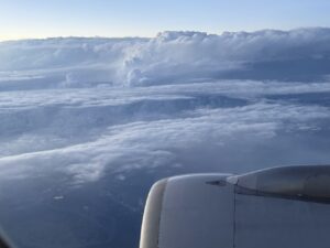 The image shows a view from an airplane window, capturing the aircraft's engine and a vast expanse of clouds below. The sky is clear with a light blue hue, and the clouds appear thick and fluffy, creating a layered effect. The horizon is visible in the distance.