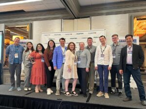 A group of eleven people is standing on a stage in front of a backdrop with logos that read "CardCon" and "Frontier." They are smiling and wearing conference badges. The setting appears to be a conference or event room with mirrors and chandeliers visible in the background.
