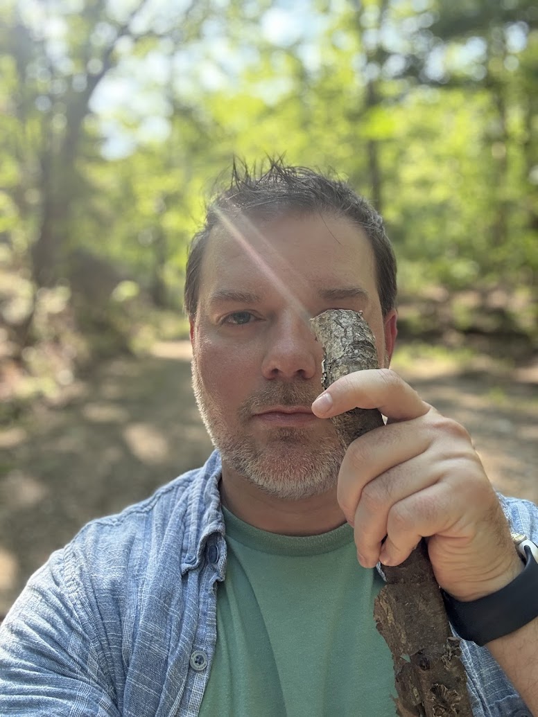 A person is standing on a forest path, holding a stick vertically in front of one eye. Sunlight filters through the trees, creating a bright and natural background. The person is wearing a light blue shirt over a green t-shirt and has a watch on their wrist.
