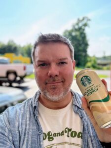 A person is holding a can of Starbucks Doubleshot Energy drink. They are wearing a striped shirt over a T-shirt with text on it. The background shows a blurred outdoor scene with trees and vehicles.