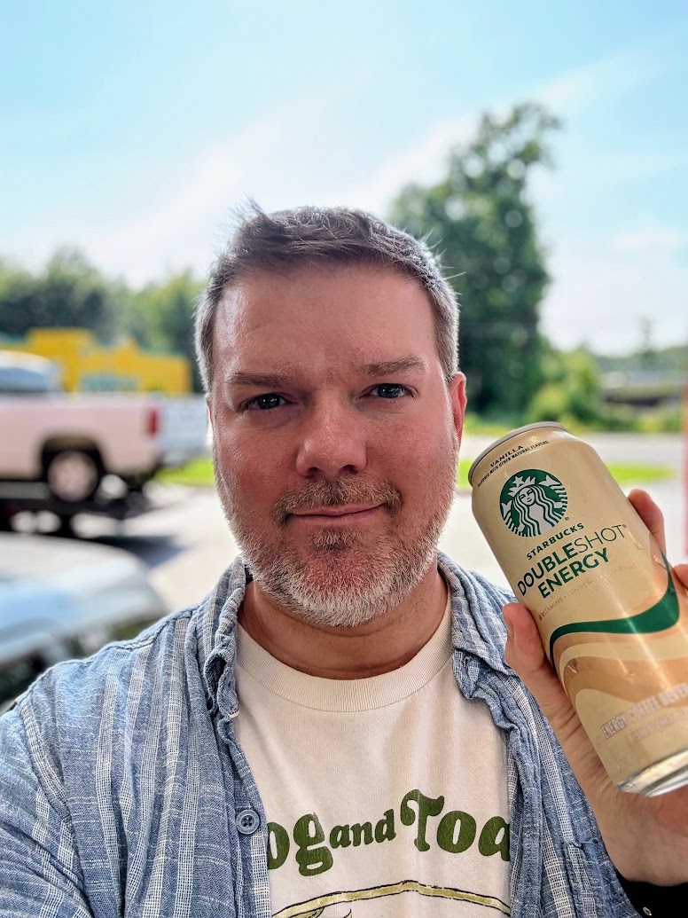 A person is holding a can of Starbucks Doubleshot Energy drink. They are wearing a striped shirt over a T-shirt with text on it. The background shows a blurred outdoor scene with trees and vehicles.