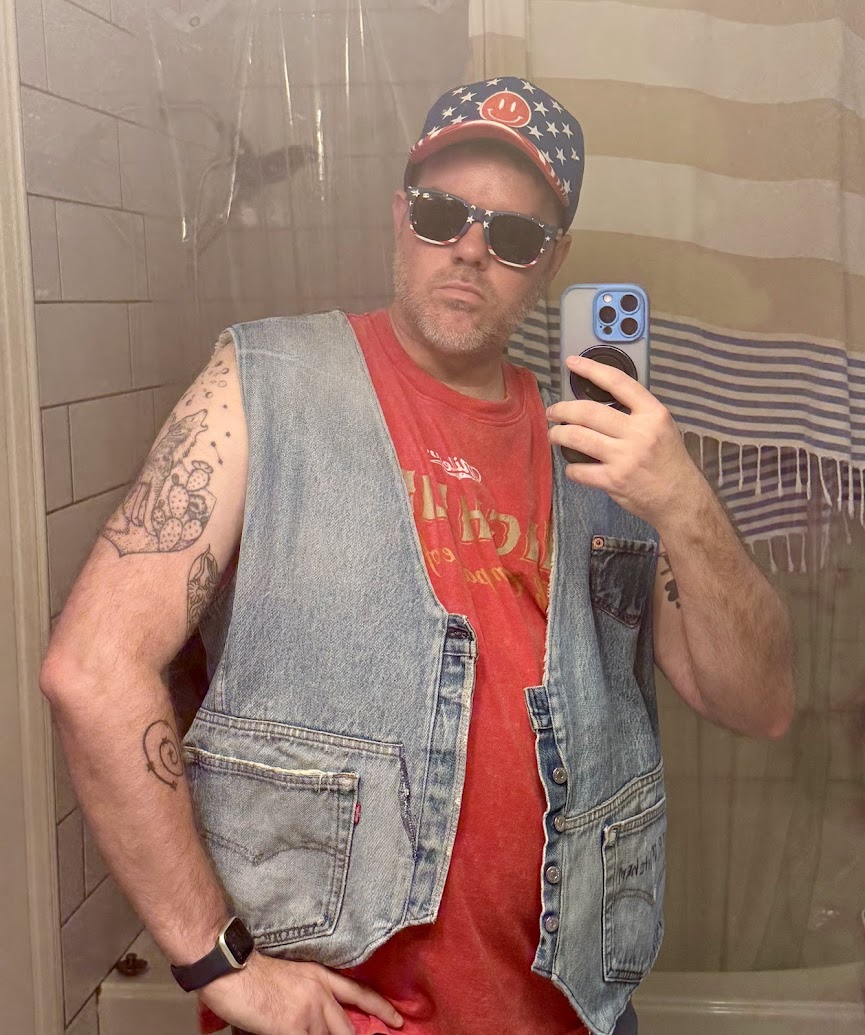 A person is taking a mirror selfie in a bathroom. They are wearing a red T-shirt, a denim vest, sunglasses, and a cap with a smiley face and star pattern. The person has tattoos on their arms and is holding a smartphone with a blue case.