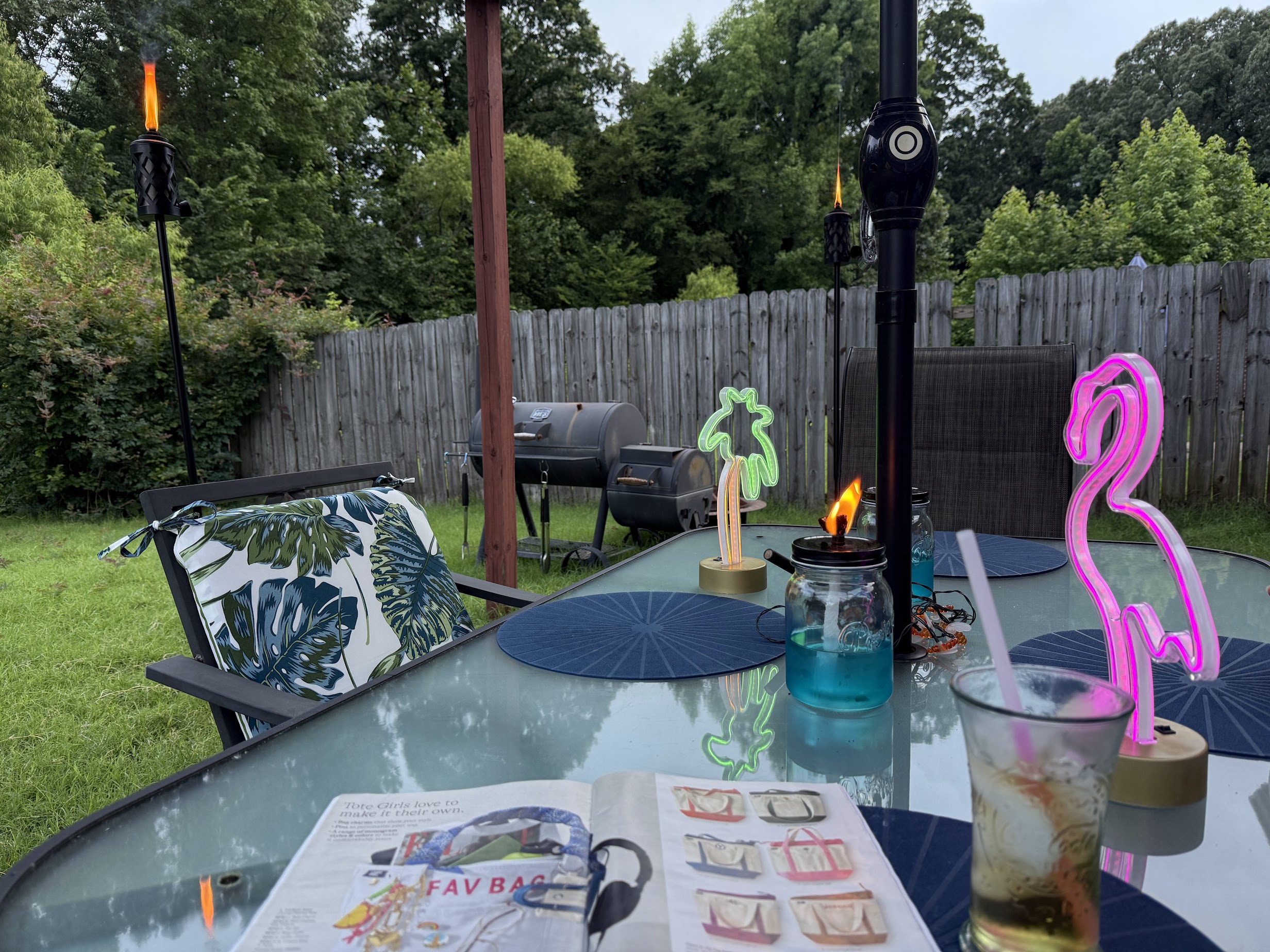 A backyard scene with a glass patio table featuring a magazine, a drink with a straw, and decorative items including a pink neon flamingo and a green neon palm tree. There are tiki torches with flames around the yard, a grill in the background, and a chair with a cushion featuring a leaf pattern. The area is enclosed by a wooden fence, and there are trees and greenery in the background.