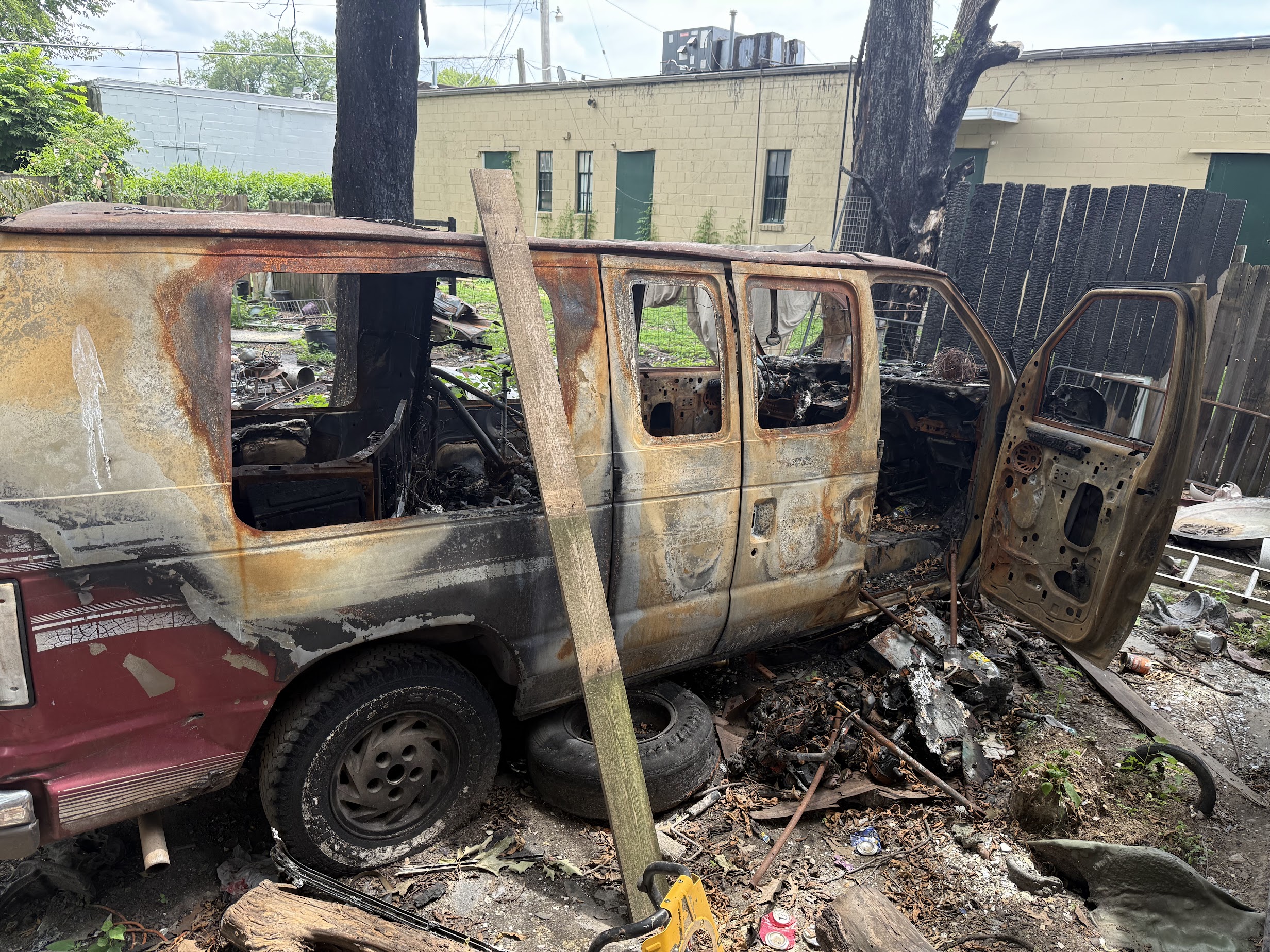 A heavily damaged and rusted van with missing windows and doors is parked in a yard. The interior is charred, indicating it has been burned. Debris and burnt materials are scattered around the vehicle. A wooden plank leans against the van, and a tree and fence are visible in the background.