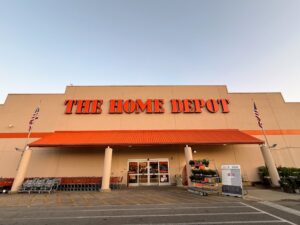 The image shows the exterior of a Home Depot store. The building has a beige facade with a large orange sign displaying "The Home Depot" at the top. There are two American flags on poles flanking the entrance. Shopping carts are lined up on the left side, and there are plants and promotional signs near the entrance. The sky is clear and blue.