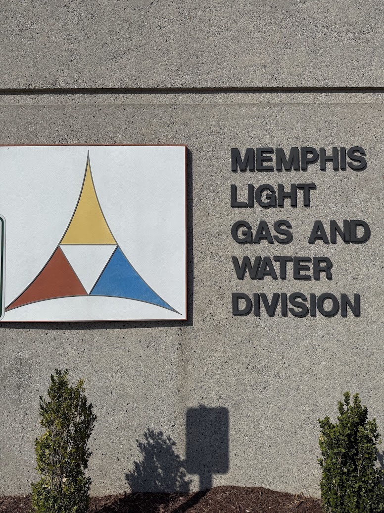 The image shows a concrete wall with the text "MEMPHIS LIGHT GAS AND WATER DIVISION" in black letters. To the left, there is a logo consisting of a triangular design with sections in yellow, red, and blue. Below the wall, there are small green shrubs planted in the soil.